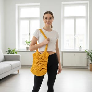 May include: A woman wearing a white t-shirt and black leggings, holding a yellow crocheted tote bag. The bag has a long strap and a net-like design. The background is a bright, modern living space with a gray sofa and plants.