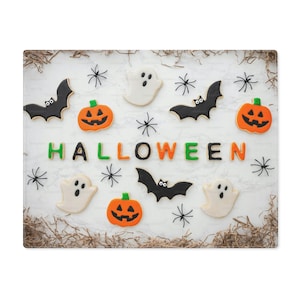 May include: A festive Halloween-themed arrangement featuring cookies shaped like bats, pumpkins, ghosts, and spiders. The word "HALLOWEEN" is spelled out in colorful cookie letters. The background is white with decorative accents.