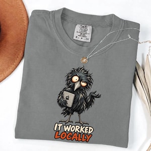 Peut inclure: Un t-shirt gris à manches courtes orné d'un dessin d'oiseau noir ébouriffé tenant une tablette argentée. Sous l'oiseau, le texte IT WORKED LOCALLY est écrit en lettres grasses orange et blanches. Le t-shirt porte une étiquette Comfort Colors.