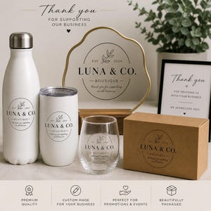May include: White water bottle, tumbler, and stemless wine glass with the "LUNA & CO. Boutique" logo. A gold-rimmed sign and a framed "Thank you" sign are also visible. A brown box with the same logo is also present.