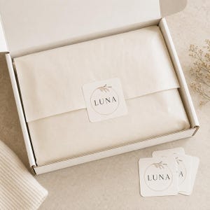 Puede incluir: Una caja de envío de cartón blanco que contiene artículos envueltos en papel de seda color crema, sellada con una pegatina cuadrada con un círculo fino, una ilustración de una ramita y el texto LUNA en fuente serif negra.