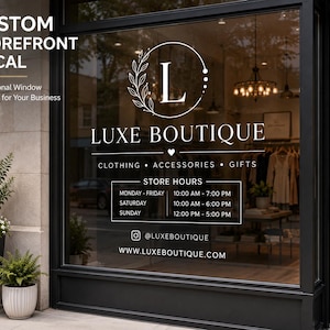 Op de afbeelding: Een witte vinyl etalagesticker op een glazen raam met de tekst LUXE BOUTIQUE, een cirkelvormig logo met een L, openingstijden en website. Het ontwerp bevat een bladmotief, een klein hartsymbool en een social media accountnaam.