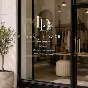 Op de afbeelding: Een glazen winkelpui met het witte logo van Lovely Daze Boutique, inclusief de tekst EST. 2022, een Instagram-account en een websiteadres. Links van de zwart omlijste glazen ingang staat een olijfboom in een pot.