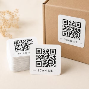 May include: A stack of square white stickers and a single square white sticker, each featuring a black QR code and the text "SCAN ME" printed in black. One sticker is applied to the side of a brown cardboard box, while others sit nearby.