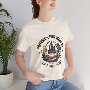 Könnte beinhalten: Cremefarbenes T-Shirt mit dem Text "Homesick for Worlds That Don't Exist". Das Design zeigt ein Schloss, ein offenes Buch, einen Mond und florale Akzente in Schwarz, Gold und Lila.