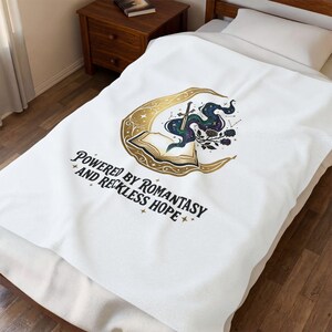 Könnte beinhalten: Weiße Decke mit einem gold-schwarzen Design mit einem Halbmond, einem offenen Buch und einem Schwert. Der Text "POWERED BY ROMANTASY AND RECKLESS HOPE" ist darunter gedruckt. Die Decke liegt auf einem Bett.