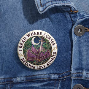Könnte beinhalten: Ein runder, weißer Emaille-Pin mit dem Text "I READ WHERE CURSES BECOME LOVE STORIES". Der Pin zeigt ein lila offenes Buch, ein Schwert, einen Mond und grüne Rosen.