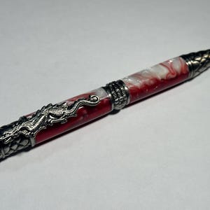 May include: A decorative pen with a red and white marbled barrel. The pen has intricate silver-coloured metalwork, including a dragon motif and textured details. The tip and end cap have a detailed, pointed design.