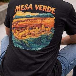 May include: Black t-shirt featuring a colorful graphic of Mesa Verde National Park with the text "MESA VERDE" in orange. The design depicts the ancient cliff dwellings and surrounding landscape in shades of orange, yellow, and blue.