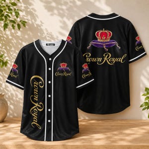 Pode incluir: Uma camisola de basebol preta com acabamentos brancos, apresentando o logótipo Crown Royal e um gráfico de uma coroa sobre uma almofada roxa. A frente exibe o texto Crown Royal verticalmente em dourado, enquanto as costas têm uma versão maior.