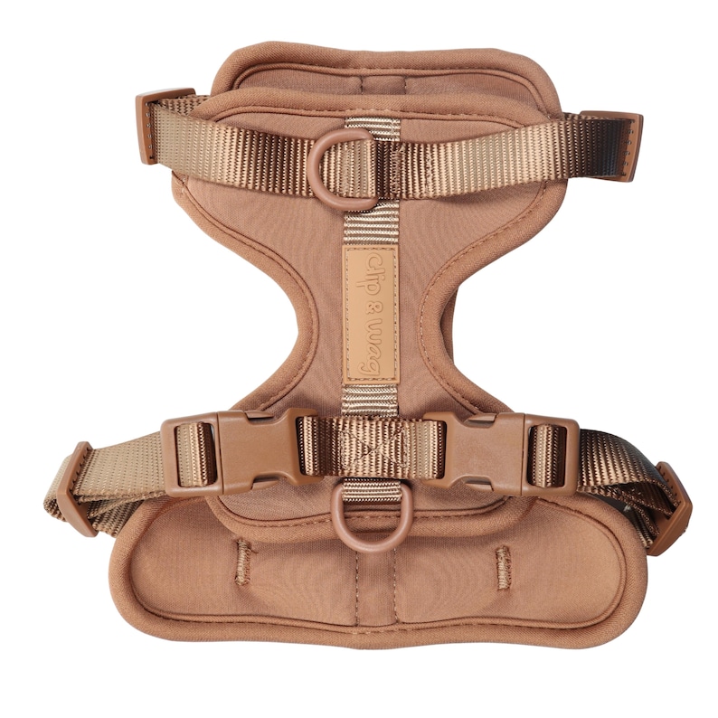 May include: A tan dog harness with adjustable straps and plastic buckles. The harness has a padded chest and back area for comfort. The brand name "clip & wag" is printed on a small tag.