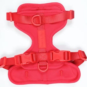 May include: A vibrant red dog harness with adjustable straps and plastic buckles. The harness has a padded chest and back area for comfort, with a D-ring for leash attachment. The brand name "elly & wumz" is printed on the strap.
