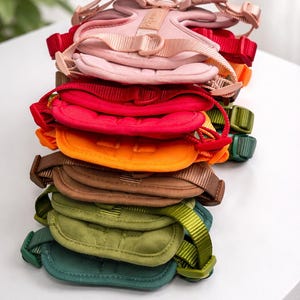 Op de afbeelding: Een stapel kleurrijke hondentuigjes in verschillende tinten, waaronder roze, rood, oranje, bruin en groen. Elk tuigje heeft verstelbare bandjes en een gewatteerde borstplaat. De tuigjes zijn netjes gerangschikt en tonen hun verschillende kleuren.