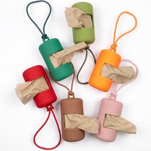 May include: A selection of brightly coloured pet waste bag dispensers in red, green, orange, brown, and pink. Each dispenser has a loop for attachment and a brown paper bag protruding. The dispensers are arranged on a white surface.