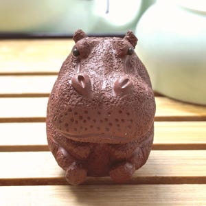 May include: A brown ceramic hippo figurine with a textured surface. The hippo has small ears, a wide snout, and tiny feet. The figurine is sitting on a wooden surface.