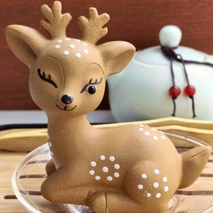 May include: A ceramic deer figurine with white spots and small antlers. The deer is light brown and sits on a clear glass plate. The deer has closed eyes and a smiling expression. A white teapot is in the background.