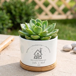 May include: A white ceramic planter with a bamboo base containing a green succulent. The planter features a black line drawing of a house with the text New Home, New Beginning, New Memories, 265 Coney Island Ave, Dahlia & Mark, and 2025.