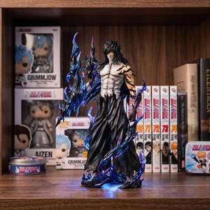 Op de afbeelding: Gedetailleerd beeldje van een personage uit de anime-serie Bleach, met een gespierd lichaam, lang zwart haar en een vloeiende zwarte outfit. Het beeldje is geaccentueerd met blauwe en paarse energie-effecten. De achtergrond bevat manga-volumes en karakterdozen.