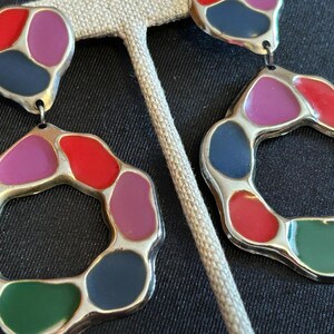 May include: A pair of colorful earrings with a silver-tone metal frame. Each earring features a unique design with red, purple, green, and blue enamel-filled shapes. The earrings are displayed on a neutral-colored earring stand.