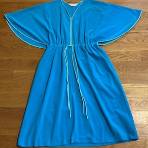 Op de afbeelding: Een turquoise jurk met een vloeiend ontwerp. De jurk heeft wijde mouwen met witte bies, een taille met trekkoord en een ritssluiting aan de voorkant. De jurk is gemaakt van een zachte, vloeiende stof.