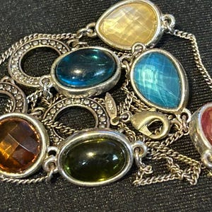 May include: A silver chain necklace with multiple colorful gemstone pendants. The stones are in various shapes and sizes, including round, oval, and teardrop. The necklace features a silver clasp.