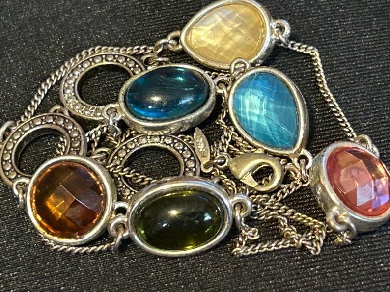 May include: A silver chain necklace with multiple colorful gemstone pendants. The stones are in various shapes and sizes, including round, oval, and teardrop. The necklace features a silver clasp.