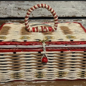 May include: A rectangular woven basket with a lid and a top handle. The basket features a pattern of cream, brown, and metallic gold woven strands. It has a red trim along the lid edge and a small white cord closure with a single red bead.