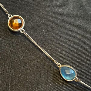 May include: A silver chain necklace with a round amber-colored gemstone and a teardrop-shaped blue gemstone.