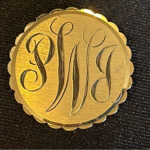 Puede incluir: Un colgante redondo de color dorado con un borde festoneado. El colgante presenta un monograma de las letras "PWG" en escritura cursiva.