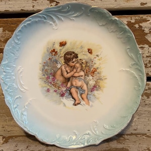 May include: A decorative porcelain plate with a light blue scalloped edge and a central image of two cherubs. The cherubs are surrounded by flowers in shades of red, pink, and yellow.