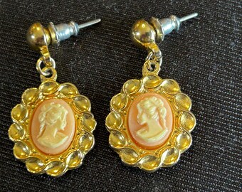 May include: A pair of gold-toned earrings with a cameo design. Each earring features a peach-colored oval cameo with a white profile, set within a scalloped gold frame. The earrings have a post-back closure.