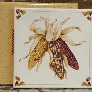 May include: A square ceramic tile featuring a detailed illustration of three ears of corn. The corn is depicted in shades of yellow, purple, and red, with tan husks. The tile has a cream-colored background and decorative corner accents.