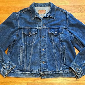 May include: A classic blue denim jacket with a button-up front, two chest pockets, and two side pockets. The jacket features a collared neckline and buttoned cuffs. A small red tag is visible on the chest pocket. The jacket is made of durable denim.