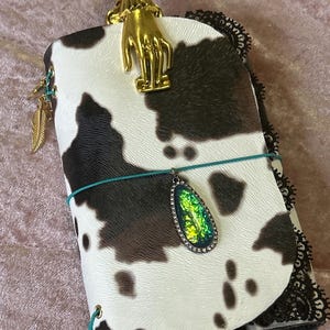 May include: A journal with a cowhide pattern cover, featuring a gold-colored hand-shaped clasp. The journal has a teal elastic closure and a decorative pendant with a green and yellow iridescent stone. A gold feather charm hangs from the side.
