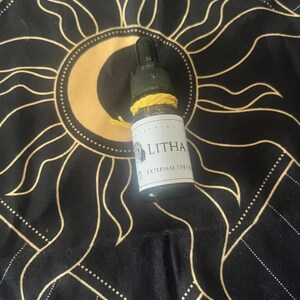 Op de afbeelding: Een kleine glazen fles met een zwarte druppelaar en een gele draad om de hals. Het etiket luidt "LITHA" en "EXTERNAL USE ONLY". De fles staat op een zwarte doek met een gouden zon- en maanontwerp.