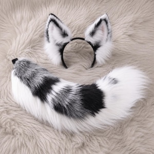 May include: A set consisting of a black headband with two pointed, faux fur animal ears and a long, curved faux fur tail. The ears and tail feature a color pattern of white, grey, and black, with long, soft, fluffy fibers throughout the design.
