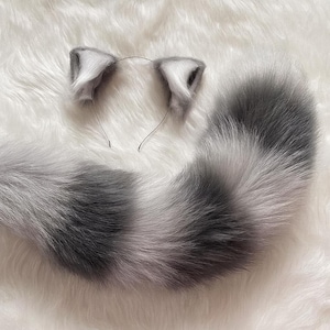 Puede incluir: Un conjunto de cola de gato y diadema de orejas de gato de piel sintética gris y blanca. Las orejas están unidas a una fina diadema de metal. La cola es larga y esponjosa, con bandas grises y blancas alternas.