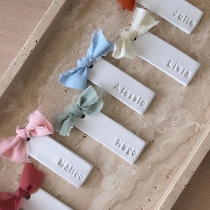 Peut inclure: Cinq étiquettes rectangulaires en argile blanche, chacune portant un nom en relief en lettres capitales : Julie, Livia, Alessio, Hugo, Matteo et Elena. Chaque étiquette est ornée d'un petit nœud en tissu effiloché, aux tons rouille, crème, bleu, sauge et rose.