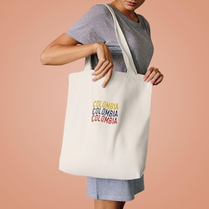 Puede incluir: Un bolso de tela color crema con la palabra COLOMBIA bordada tres veces en un diseño apilado. La palabra superior es amarilla, la del medio es azul y la inferior es roja, reflejando los colores de la bandera de Colombia.