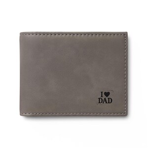 Puede incluir: Una cartera de cuero gris con el texto "I ♥ DAD" impreso en negro. La cartera es rectangular y tiene un borde cosido. La cartera es de estilo plegable.
