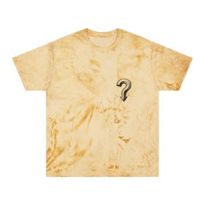 Puede incluir: Camiseta de manga corta con un estampado jaspeado marrón y beige. Una interrogación negra y plateada con una flecha está impresa en la parte delantera. La camiseta tiene cuello redondo.