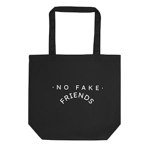 May include: Black tote bag with white text that reads "NO FAKE FRIENDS" in a curved font. The bag has two black handles and is made of a sturdy material, suitable for carrying various items. The design is simple and bold.