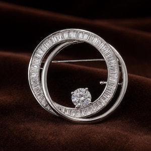 May include: A silver-toned circular brooch featuring a ring encrusted with numerous small baguette-cut clear stones. A single, larger round brilliant-cut clear stone is positioned inside the circle, attached to the inner edge of the frame.