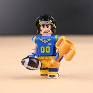 Peut inclure: Figurine de style Lego représentant un joueur de football américain en uniforme bleu et jaune, tenant un ballon de football marron et un trophée doré. La figurine porte un casque noir et le numéro "00" sur le maillot.