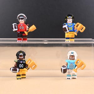 Op de afbeelding: Vier miniatuurfiguren van American footballspelers in verschillende teamkleuren. Elke figuur houdt een voetbal en een trofee vast. De figuren dragen helmen en shirts met het nummer "00" of "10". De figuren staan op een doorzichtige plank.