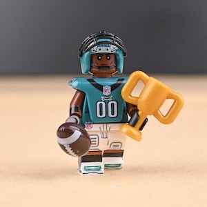 Könnte beinhalten: Miniatur-Footballspieler-Figur in einem türkisblauen Trikot mit der Nummer 00, einem schwarzen Helm und einer weißen Hose. Die Figur hält einen Football und eine goldene Trophäe. Das NFL-Logo ist sichtbar.