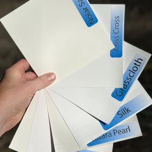May include: A hand holds a fan of white paper samples with blue labels. The labels read "Nolar Sailcloth", "Criss Cross", "Glasscloth", "Silk", "Matara Pearl", "Peel & Stick", and "Ultra Smooth". The papers have various textures.