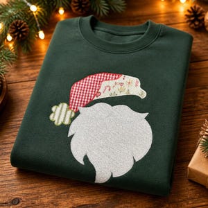 May include: A dark green crewneck sweatshirt featuring a central applique of a Santa Claus face. The design includes a white textured beard and a hat with a red and white gingham pattern, a floral print section, and a green and white striped pom-pom.
