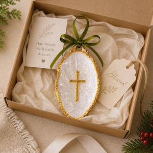 May include: An oval-shaped white resin ornament with a gold-colored cross in the center, accented by a green ribbon bow. The ornament is presented in a cardboard box with two paper tags, one reading Handmade with Faith & Love and one reading Blessed.