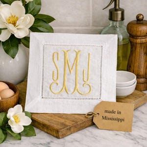 May include: White linen monogrammed napkin with gold embroidery. The napkin features the initials "SMJ" in an elegant script. A tag that says "made in Mississippi" is attached. The napkin is displayed with flowers and kitchen accessories.